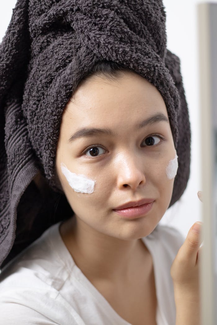 Young woman in towel wrap applying face cream, indoors, skincare routine.