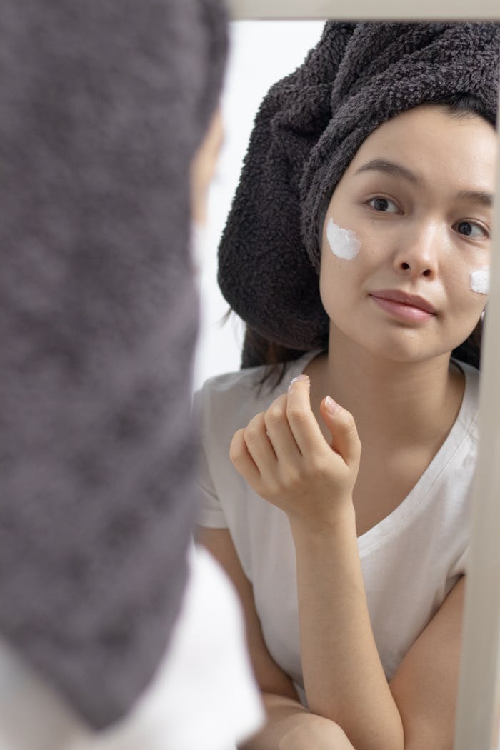 Woman in towel applying face cream, looking at her reflection. Skincare routine in the bathroom.