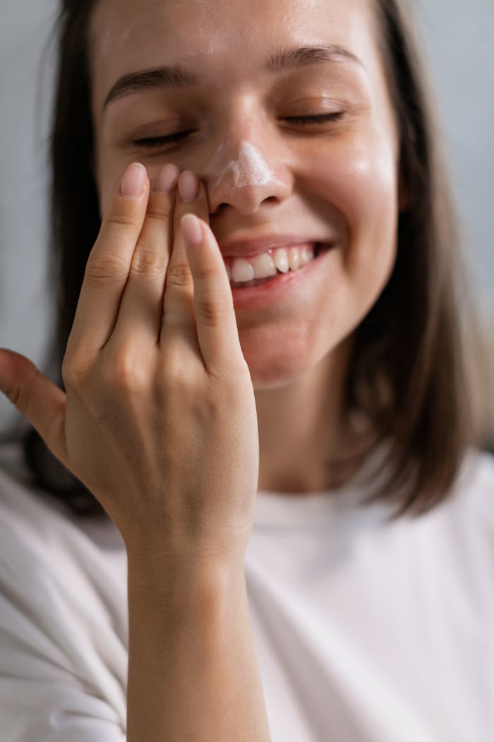 Smiling young adult woman applying skincare cream to her face in a gentle light setting, promoting beauty care.
